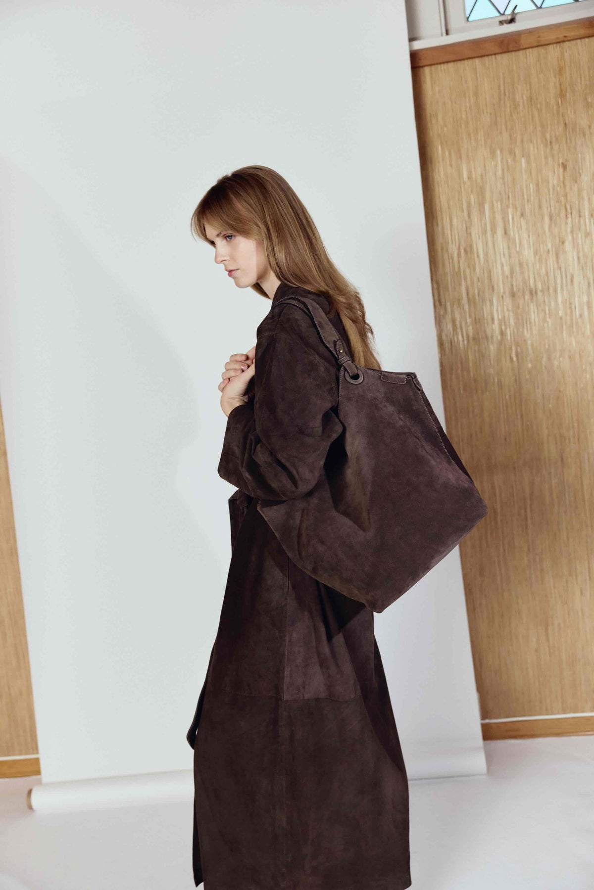 Woman wearing a long brown coat holding a matching bag indoors
